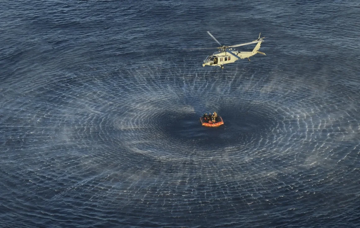 A helicopter flies above a recovery raft surrounded by wide circular ripples in the Pacific after the Artemis II splashdown.