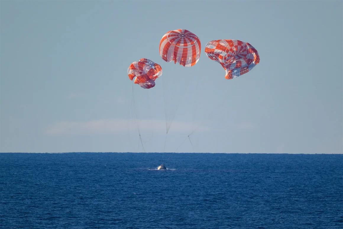 NASA’s Orion spacecraft sits low over the Pacific Ocean beneath three orange-and-white main parachutes during the Artemis II splashdown sequence.