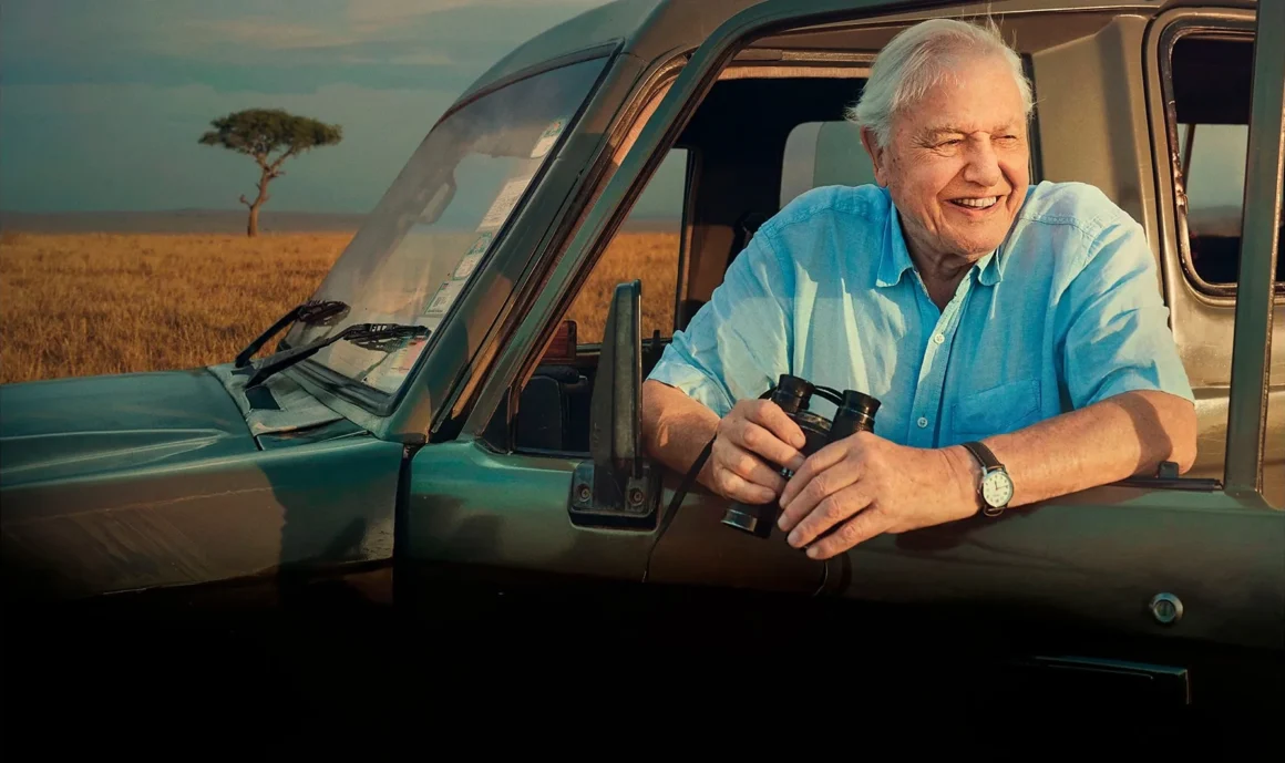 David Attenborough smiling while leaning out of a safari vehicle in Kenya, holding binoculars and looking across the savannah.