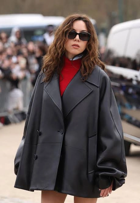Aubrey Plaza walking outdoors during Paris Fashion Week in an oversized black leather jacket and sunglasses.