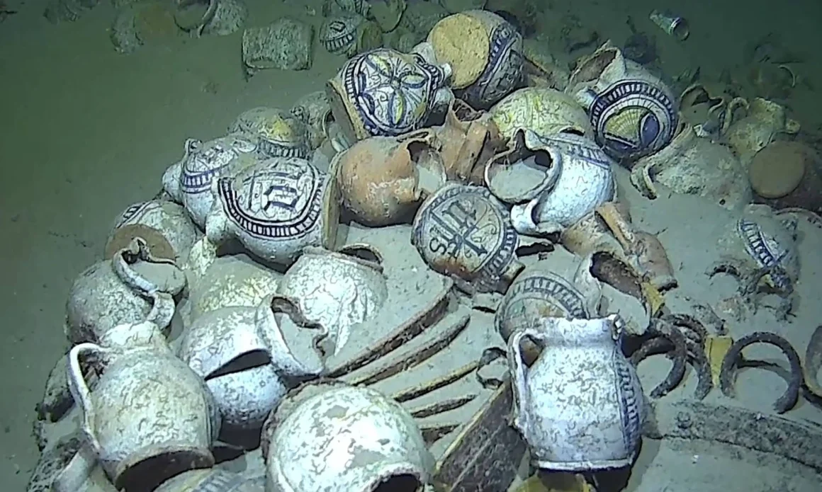 Close-up underwater view of ceramic jugs, bowls, and stacked plates from the Camarat 4 shipwreck resting on the seafloor.
