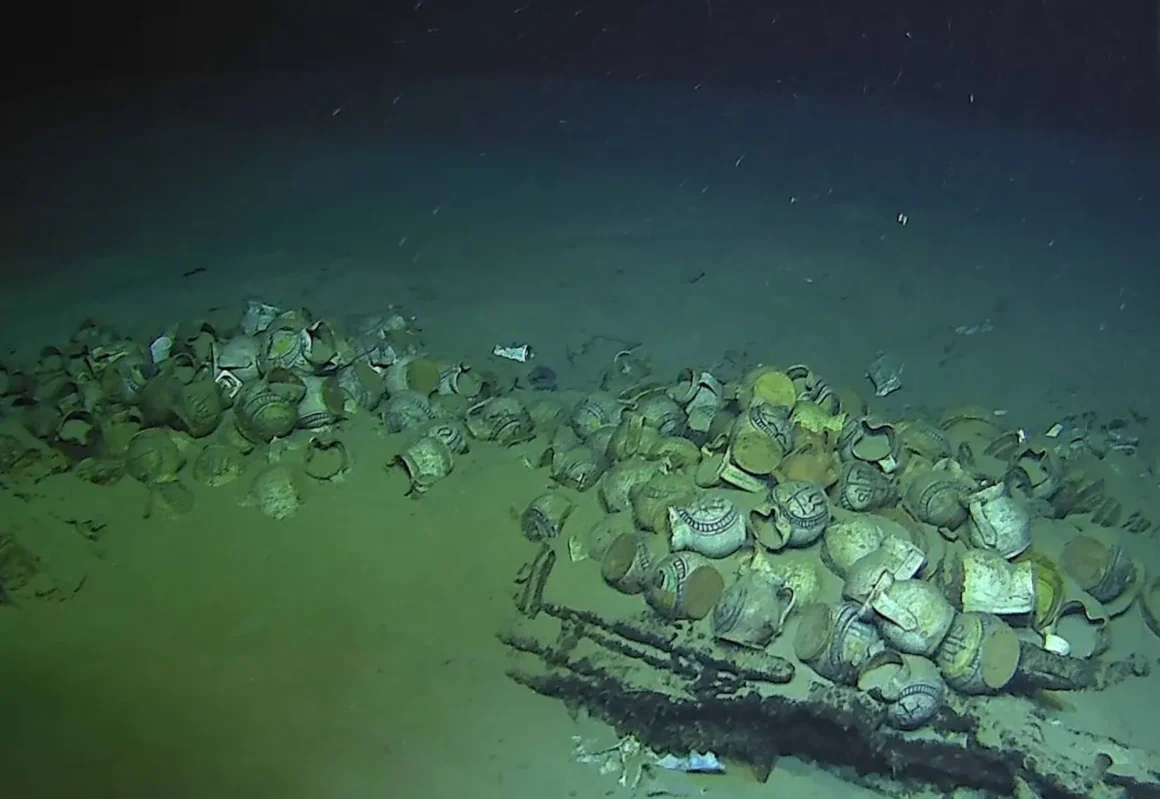 Wide underwater view of ceramic jugs and bowls scattered across the Camarat 4 shipwreck site on the seafloor.