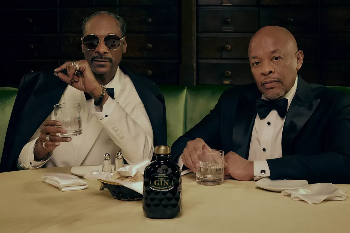 Dr. Dre and Snoop Dogg sitting in formal attire at a table with glasses and a bottle of Still G.I.N. in front of them