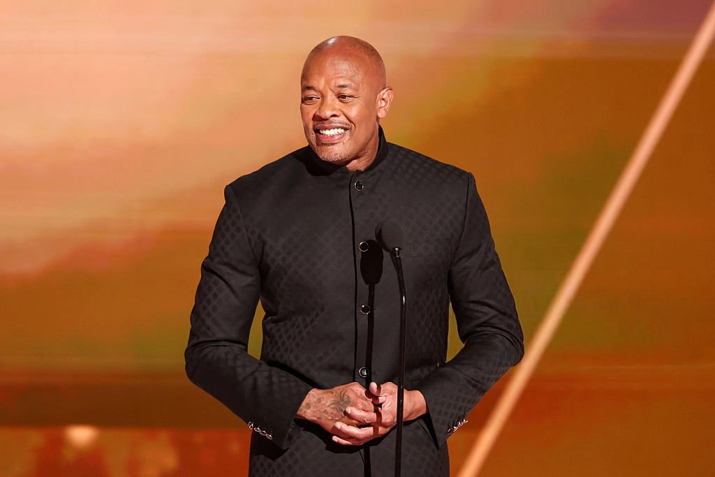 Dr. Dre standing on stage in a black outfit speaking into a microphone under warm lighting