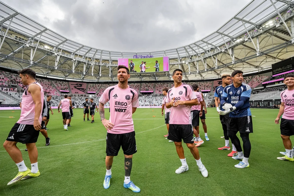 Inter Miami players stand on the pitch inside Nu Stadium before a match, with the stadium roof and central screen visible above them.