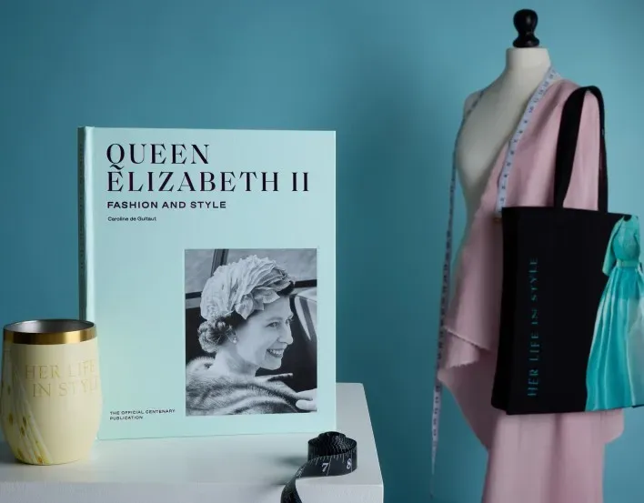 Display featuring the Queen Elizabeth II: Fashion and Style book beside a dress form with exhibition merchandise in soft blue and pink tones.