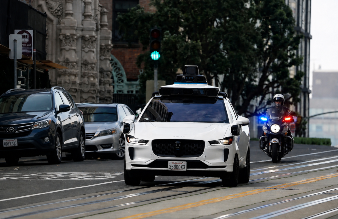 A driverless car drives through a city street while a police motorcycle follows behind with lights on